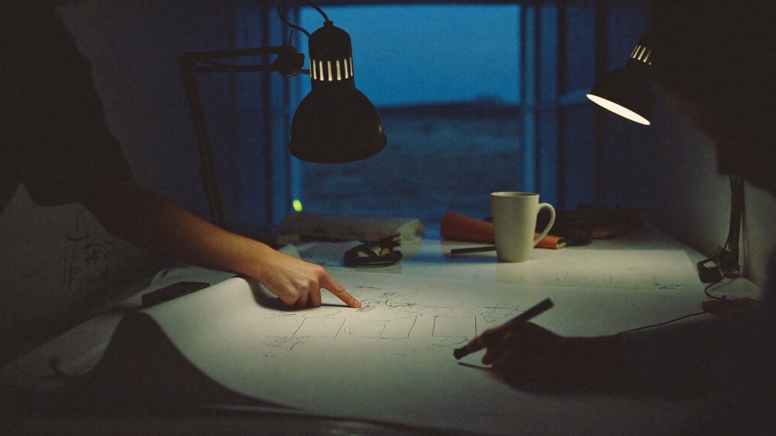 Two people pointing at construction drawings on a blueprint table in a jobsite trailer at blue hour