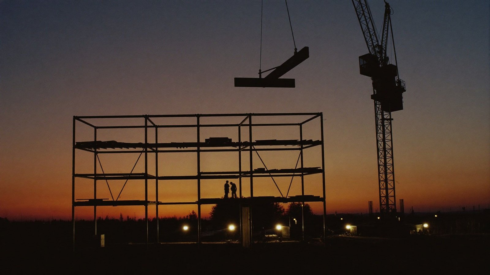 Tower crane setting a steel beam onto a building frame at dusk, two ironworkers waiting on the top level, work lights at grade