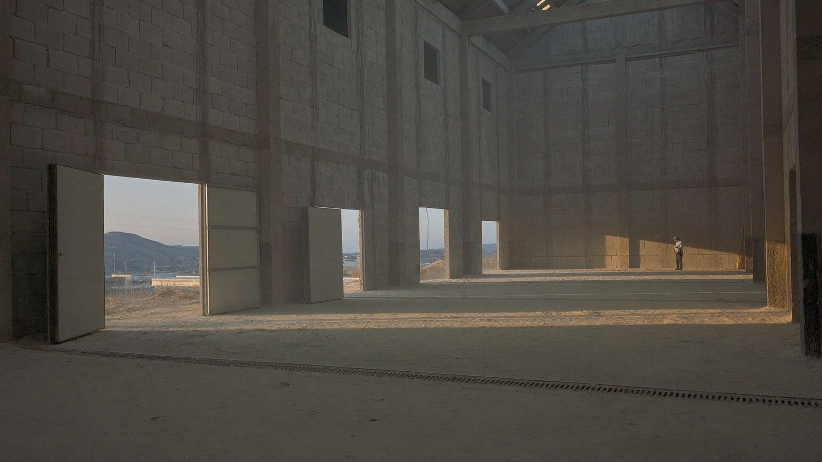 Golden-hour light raking through an open bay door of an unfinished tilt-wall warehouse, foreman reviewing work at the far end